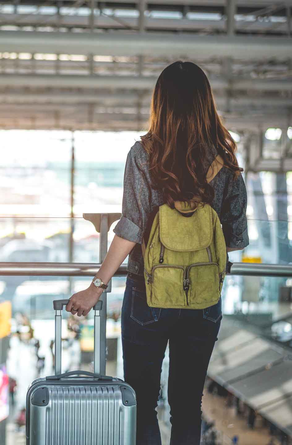Frau mit gelbem Rucksack und Koffer schaut auf Flughafen-Terrasse nach draußen