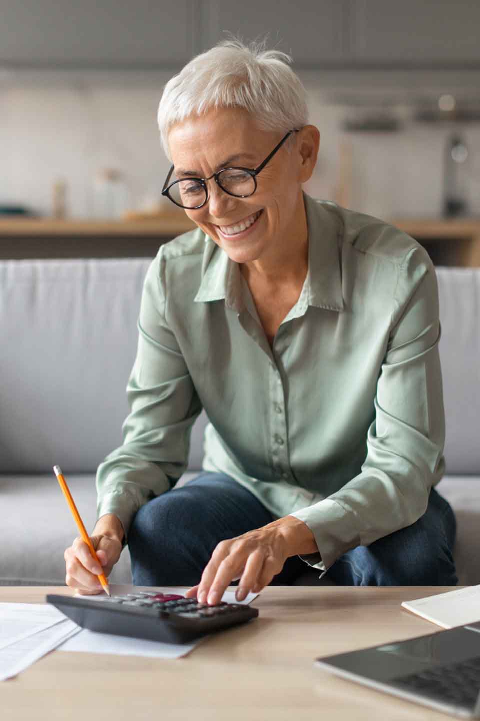 Ältere Frau mit Brille arbeitet lächelnd mit Taschenrechner und Stift