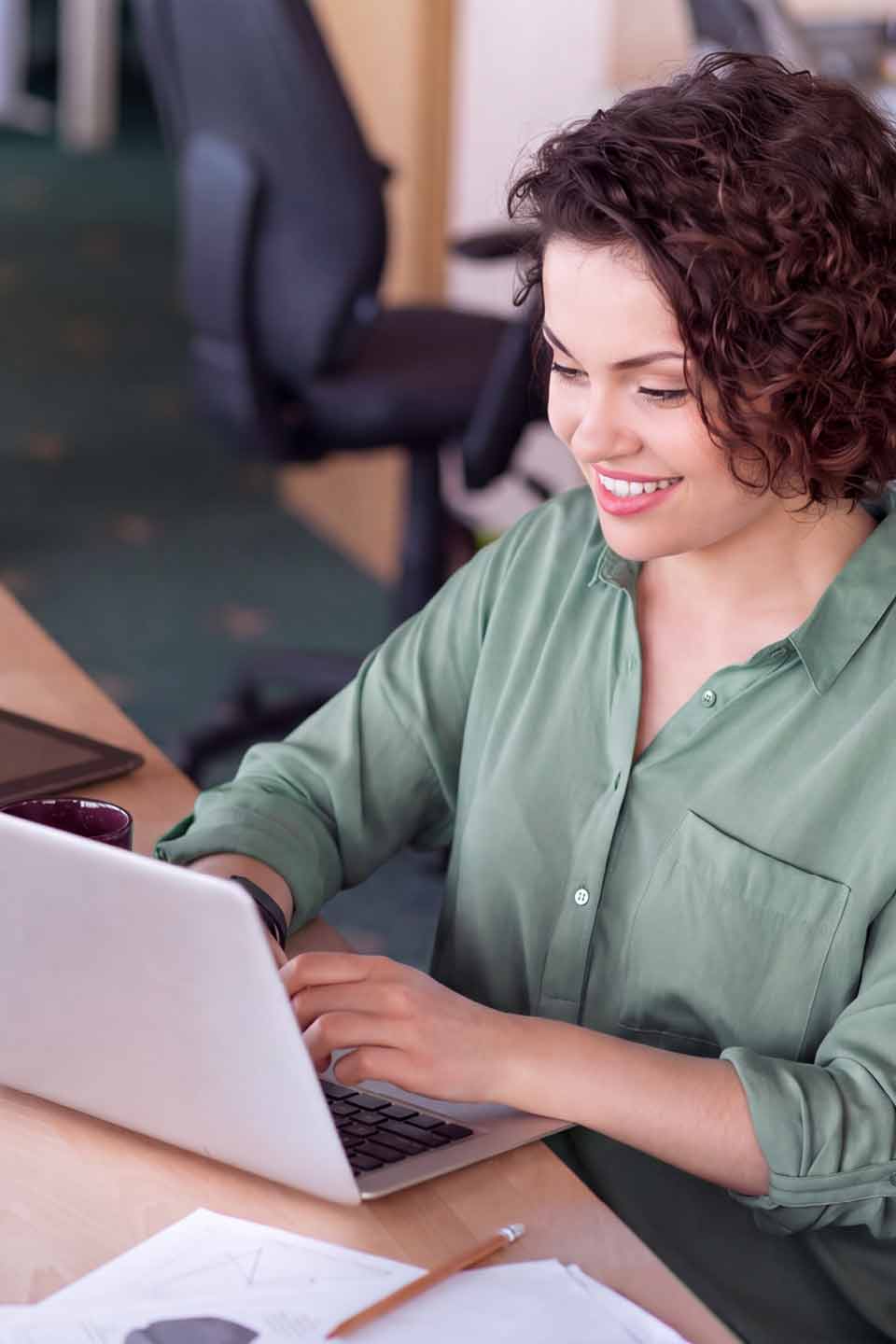 Lächelnde Frau mit lockigem braunem Haar und grünem Hemd arbeitet konzentriert an einem Laptop in einem Büro.