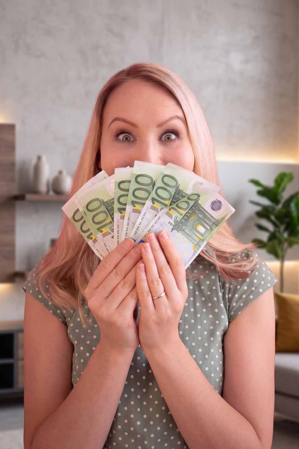 Überraschte Frau mit rosafarbenen Haaren hält mehrere 100-Euro-Scheine in den Händen und schaut erstaunt in die Kamera.