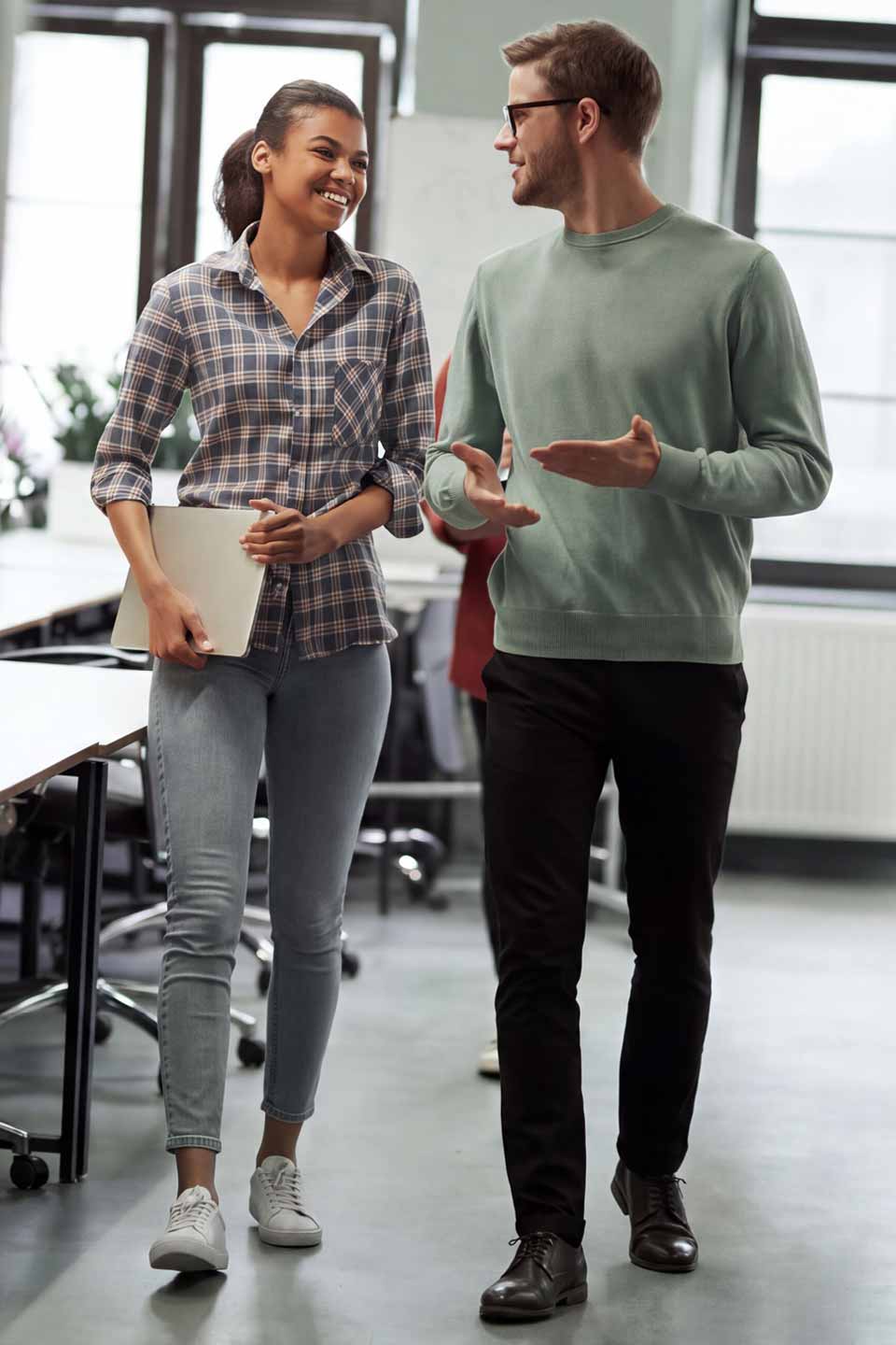 Studierende gehen zusammen durch ein modernes Büro und unterhalten sich. Die Frau trägt ein kariertes Hemd und hält einen Laptop, der Mann trägt einen grünen Pullover und eine Brille.