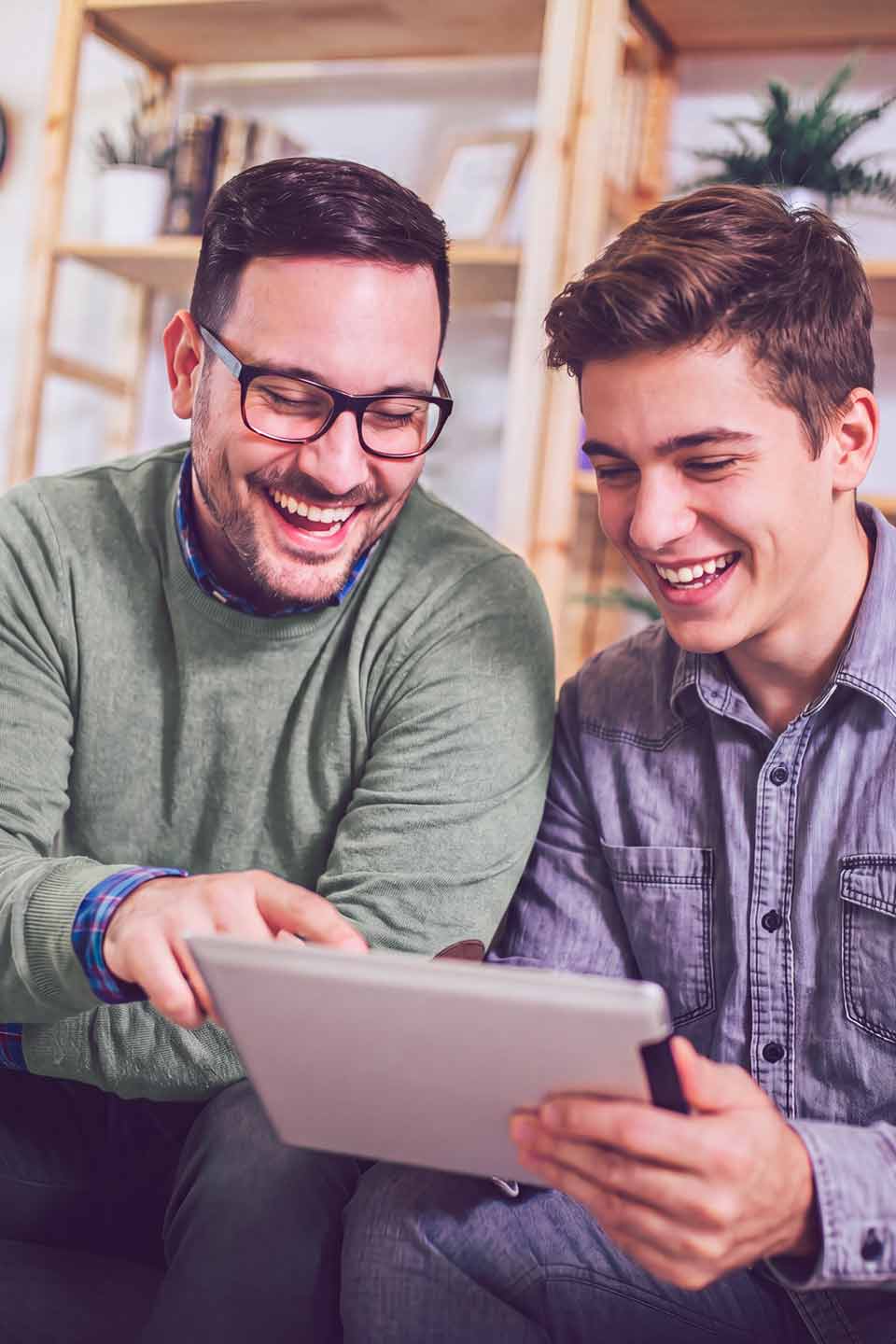 2 Studierende sitzen zusammen und schauen auf ein Tablet. Der Mann links trägt eine grüne Jacke und eine Brille, der Mann rechts trägt ein lila Jeanshemd.