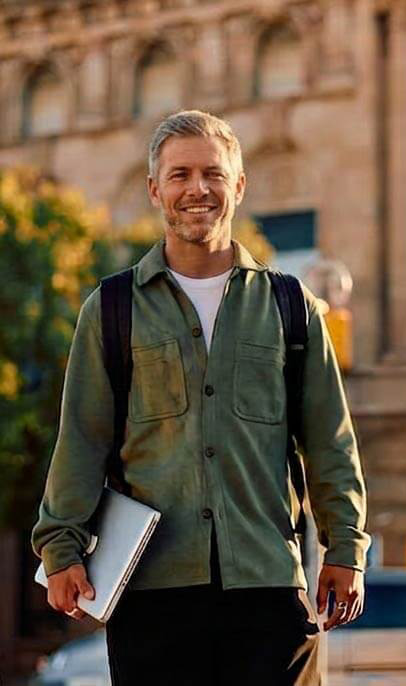 Ein lächelnder Mann mittleren Alters in olivfarbener Jacke mit Laptop steht vor historischen Gebäuden in einer sonnigen Universitätsstadt.