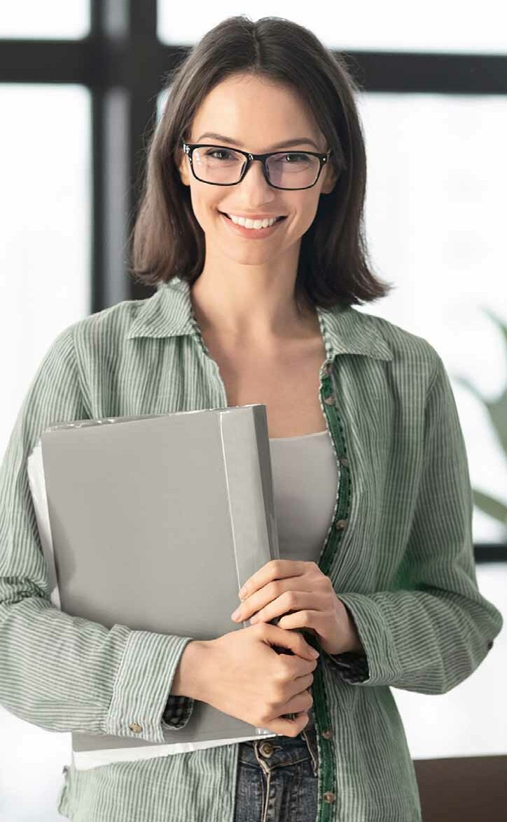 Porträt einer lächelnden Frau mit Brille, die einen Laptop hält und ein grünes Hemd trägt, vor einem modernen Büro-Hintergrund mit Fenster.